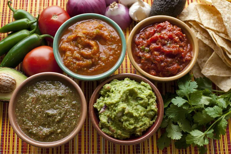 Four bowls of mexican salsas and guacamole arranged on a striped placemat with fresh ingredients around