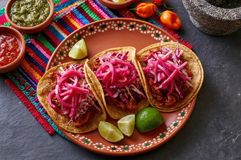 Three cochinita pibil tacos with pickled onions and lime on a hand-painted terracotta plate over a colorful woven placemat