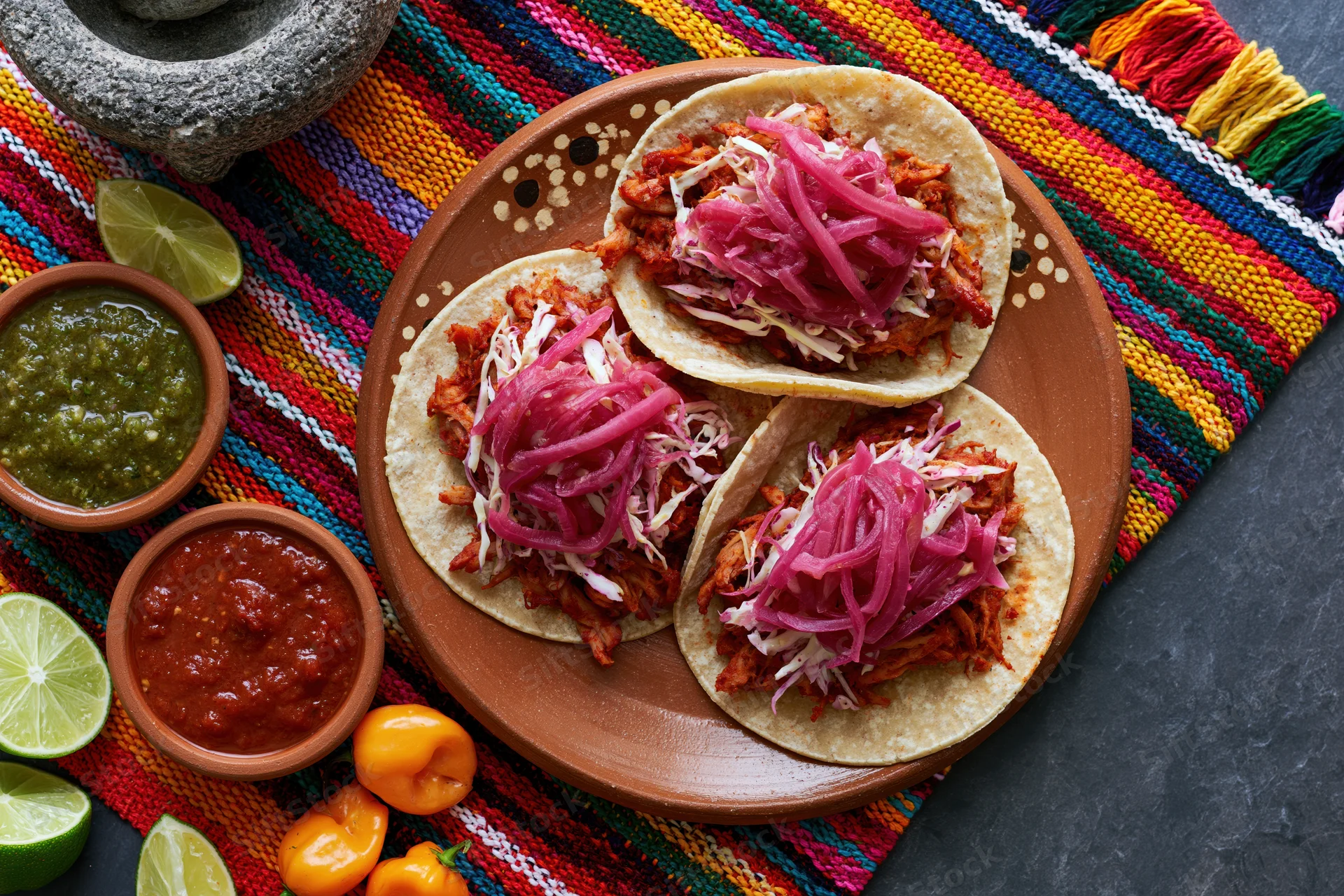 Three cochinita pibil tacos on a hand-painted terracotta plate with salsas and lime on a colorful woven placemat