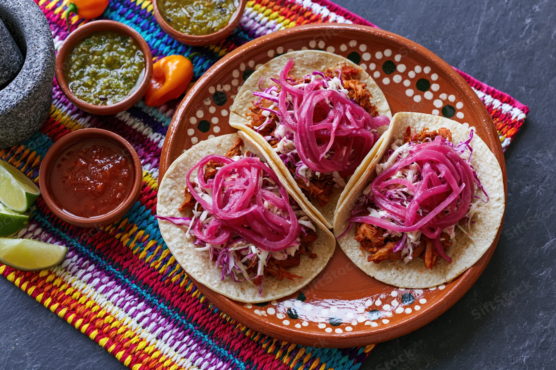 Three cochinita pibil tacos on a hand-painted terracotta plate with pickled onions and salsas on a colorful woven placemat