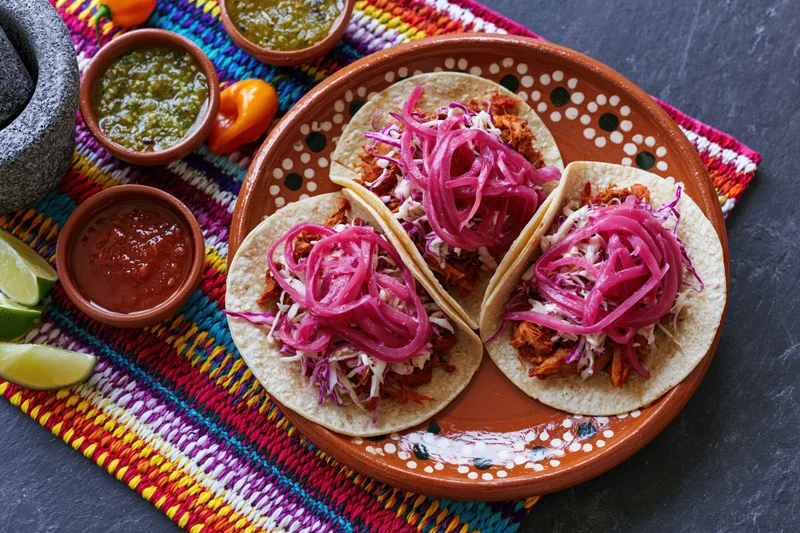 Three cochinita pibil tacos on a hand-painted terracotta plate with pickled onions and salsas on a colorful woven placemat