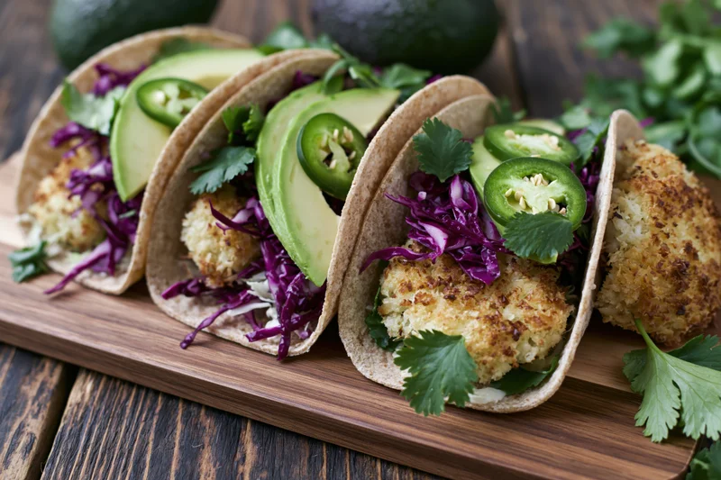 Three crispy coconut-crusted cauliflower tacos with avocado and purple cabbage on a wooden cutting board