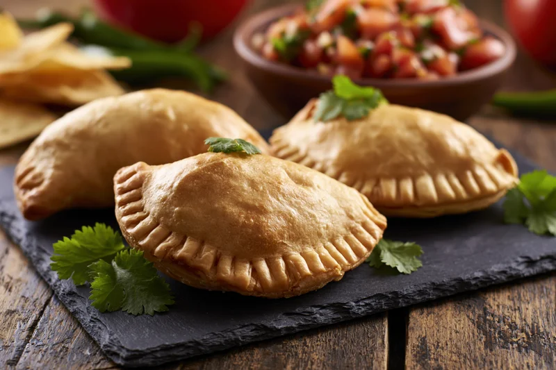 Golden-baked empanadas arranged on a dark slate board with cilantro and salsa on a wooden table