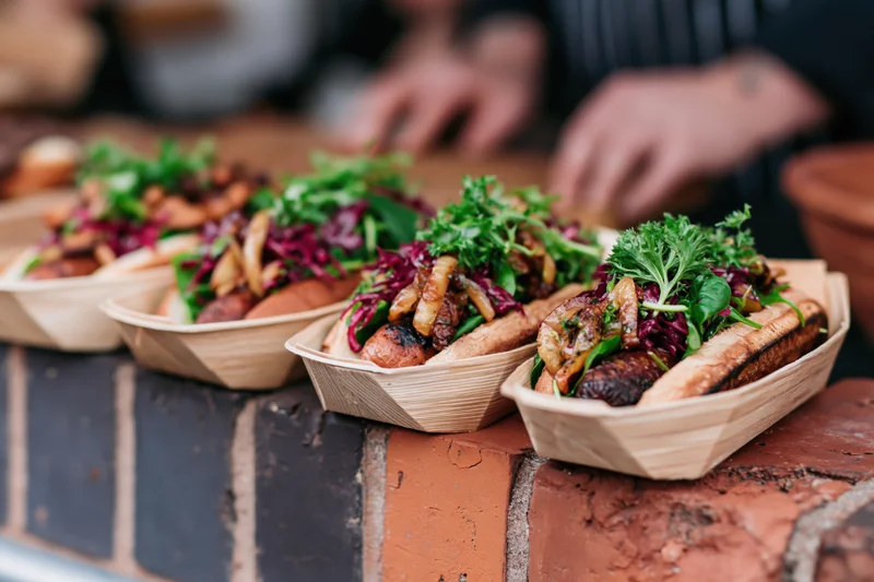 Gourmet hot dogs with vegetables and toppings served in wooden boat trays on a brick counter