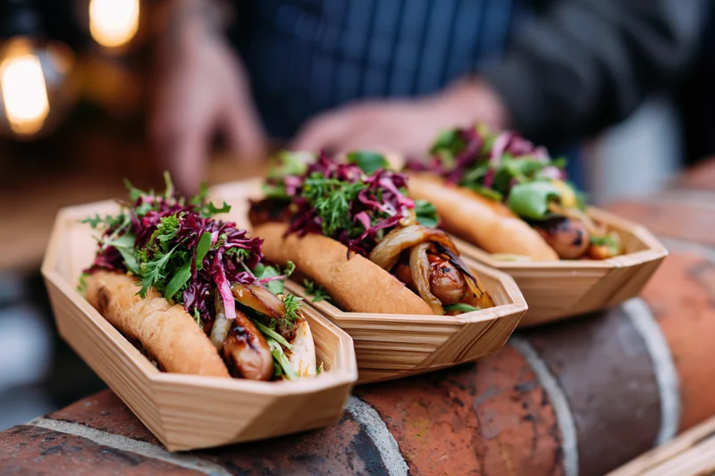Three gourmet hot dogs with fresh toppings served in wooden boat trays on a brick surface