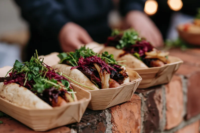 Three gourmet hot dogs with toppings in wooden boat trays lined up on a brick wall