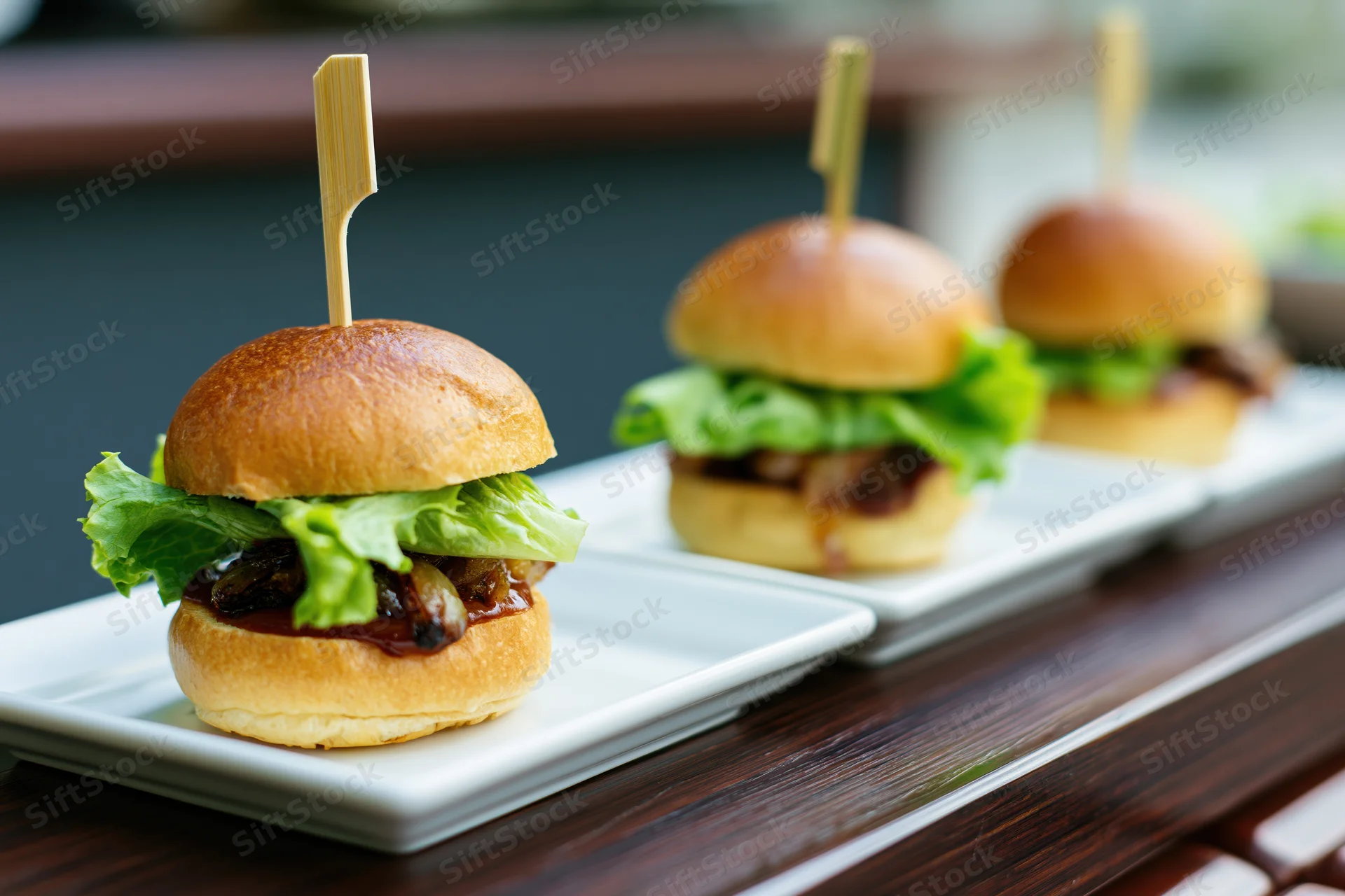 Three gourmet mini sliders on white rectangular plates arranged in a row on a wooden table
