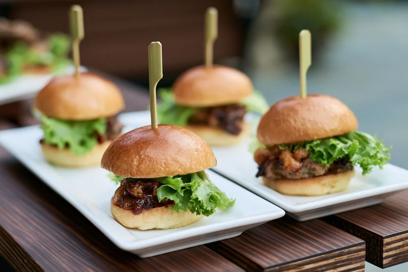 Gourmet mini sliders with lettuce and sauce on white rectangular plates arranged on a wooden table