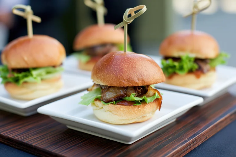 Gourmet mini sliders on white square plates arranged on a wooden tray for serving at an event