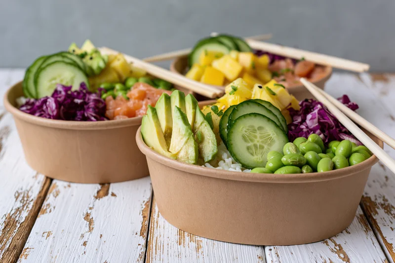 Three poke bowls with rice, avocado, cucumber, pineapple, edamame, red cabbage, and salmon in kraft paper containers on a white wooden table