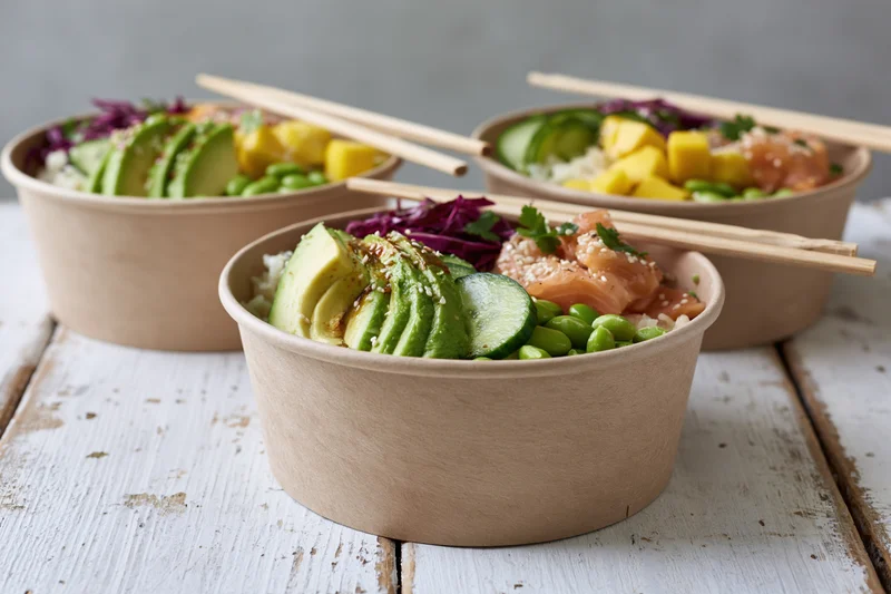 Three poke bowls with avocado salmon edamame and mango in kraft paper containers on a white wooden table