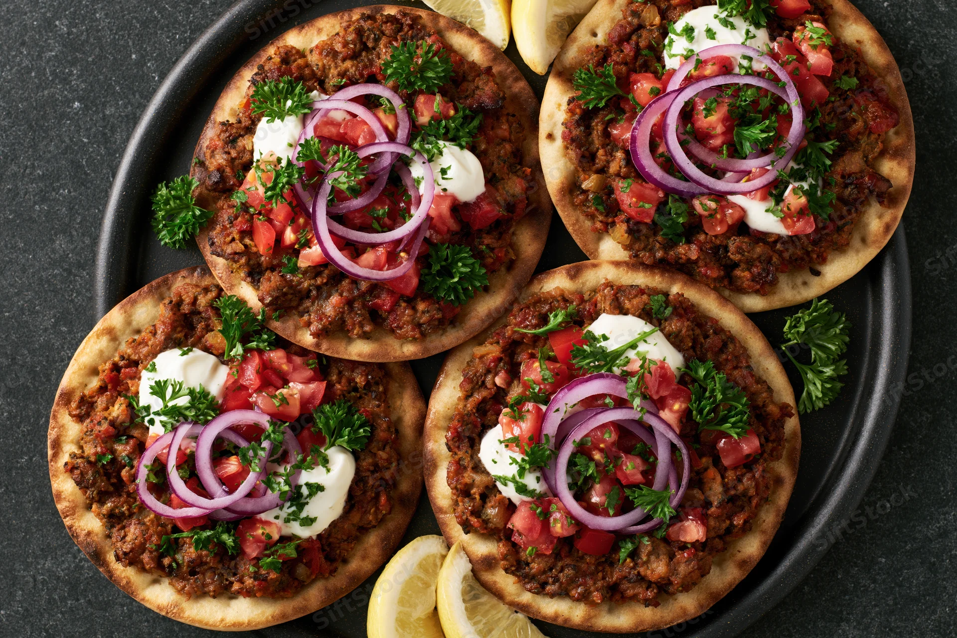 Four lahmacun flatbreads topped with spiced ground meat, red onions, tomatoes, parsley and yogurt on a black plate with lemon slices