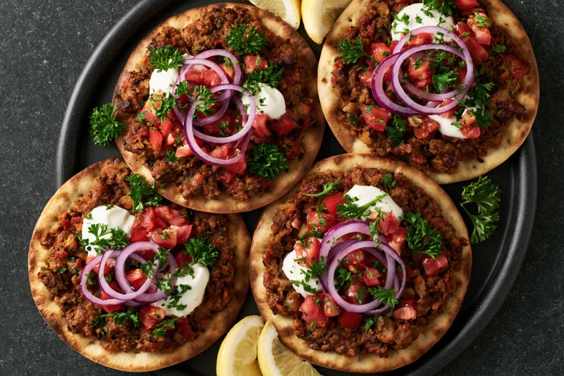 Four lahmacun flatbreads topped with spiced ground meat, red onions, tomatoes, parsley and yogurt on a black plate with lemon slices
