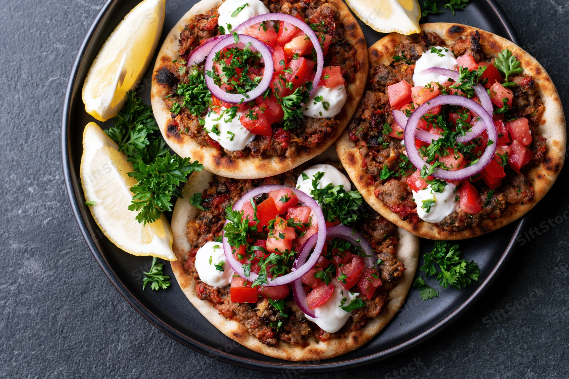 Three lahmacun flatbreads topped with spiced ground meat and fresh vegetables on a black plate