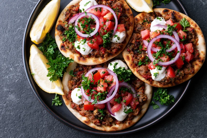 Three lahmacun flatbreads topped with spiced ground meat and fresh vegetables on a black plate