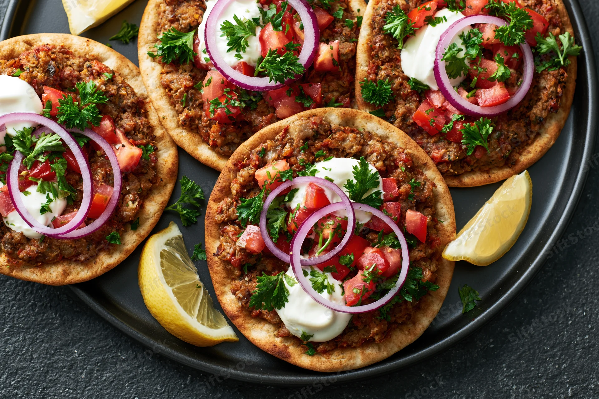 Four lahmacun flatbreads topped with spiced ground meat, tomato, onion, parsley and yogurt on a black plate with lemon wedges