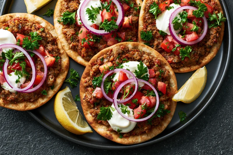 Four lahmacun flatbreads topped with spiced ground meat, tomato, onion, parsley and yogurt on a black plate with lemon wedges