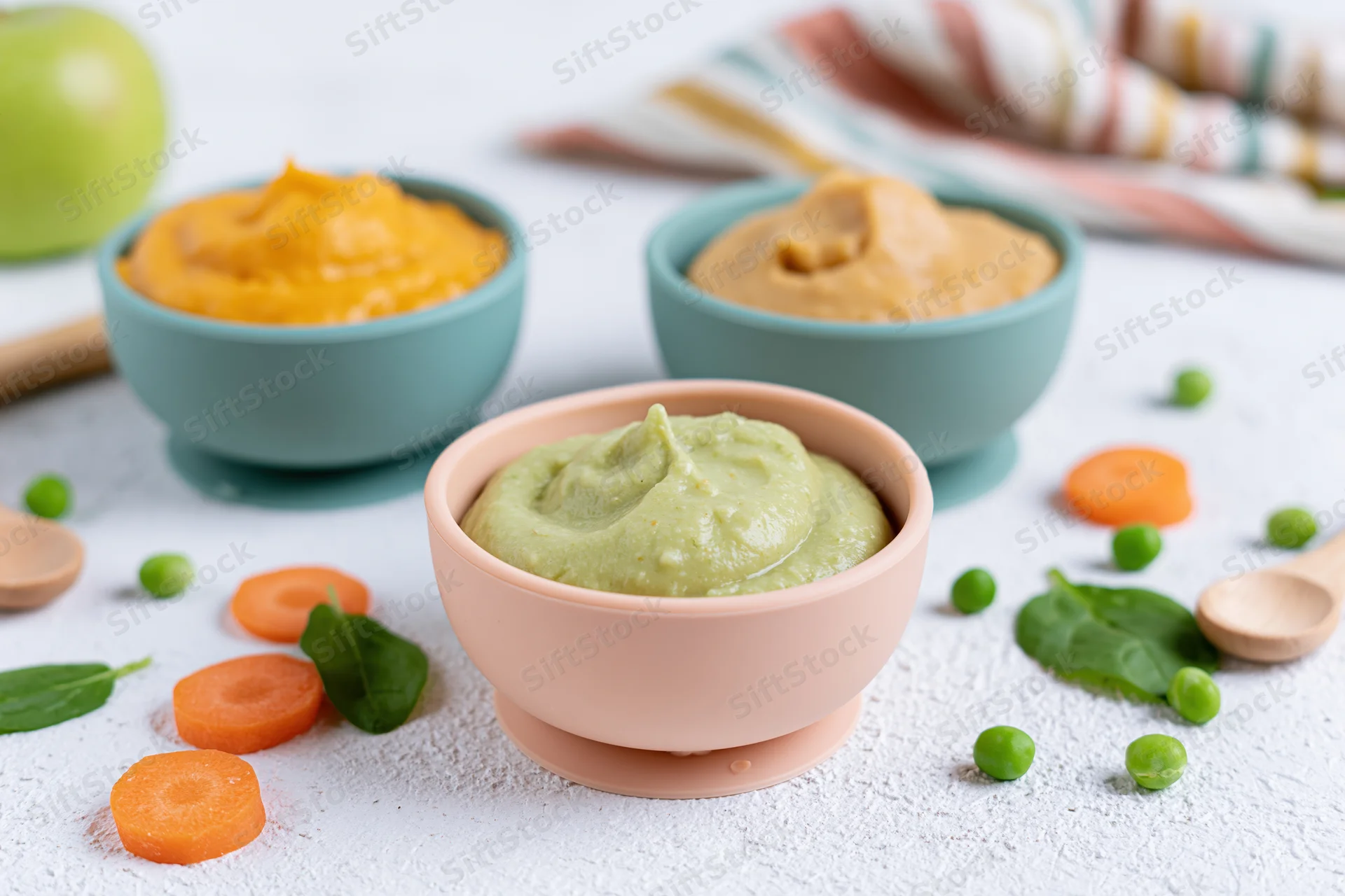 Three pastel silicone baby bowls filled with vegetable purees arranged on a white surface with scattered carrots, peas, spinach, and wooden spoons