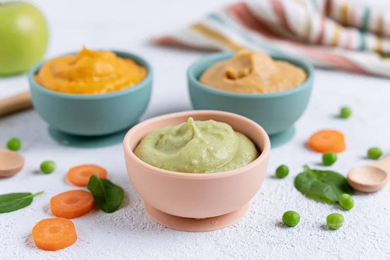 Three pastel silicone baby bowls filled with vegetable purees arranged on a white surface with scattered carrots, peas, spinach, and wooden spoons