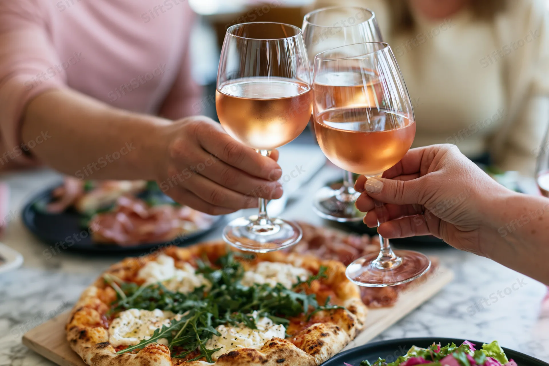 Women clinking rosé wine glasses over a shared pizza at a restaurant table