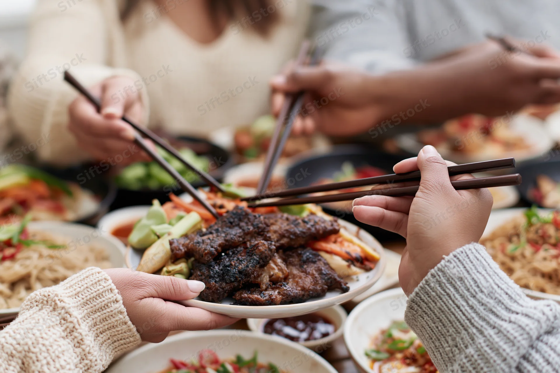 Hands using chopsticks to share grilled meat and vegetables from a plate at a communal dining table