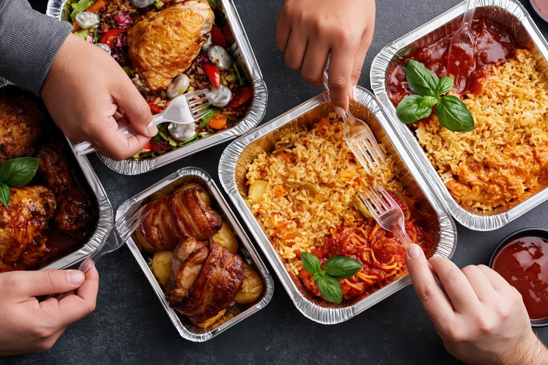 Hands reaching into aluminum takeout containers filled with various meals on a dark surface