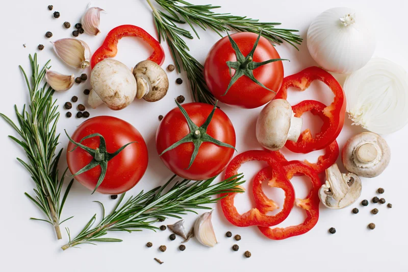 Ripe tomatoes with rosemary, garlic, onion, mushrooms and red pepper slices arranged on white surface