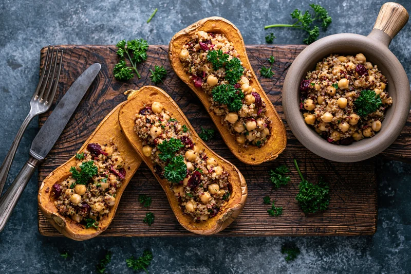 Roasted butternut squash halves stuffed with quinoa and chickpeas on a wooden board with cutlery and herbs