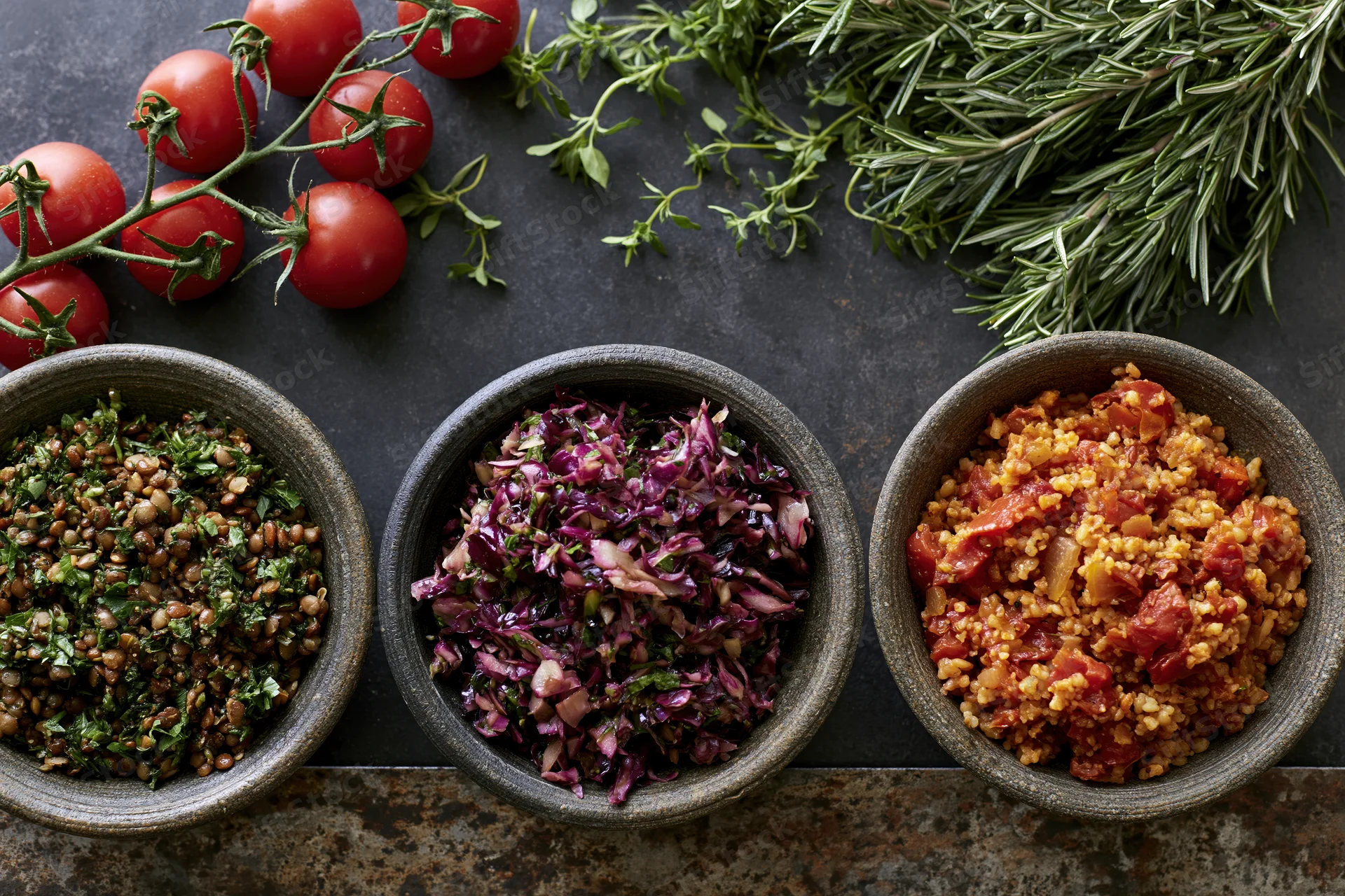 Three rustic bowls of grain and vegetable salads arranged on a dark stone countertop with fresh herbs and cherry tomatoes overhead