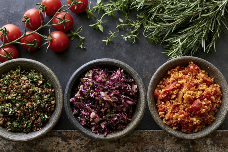 Three rustic bowls of grain and vegetable salads arranged on a dark stone countertop with fresh herbs and cherry tomatoes overhead
