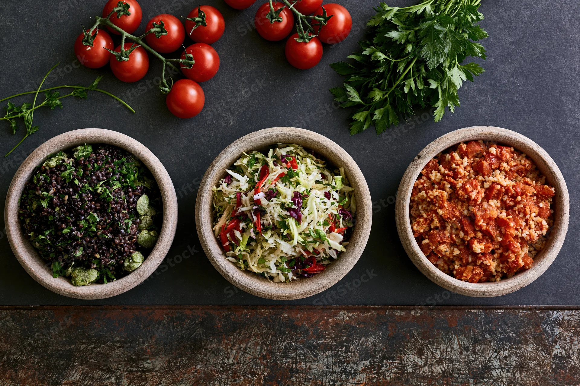 Three rustic bowls of grain and vegetable salads arranged on a dark surface with tomatoes and parsley