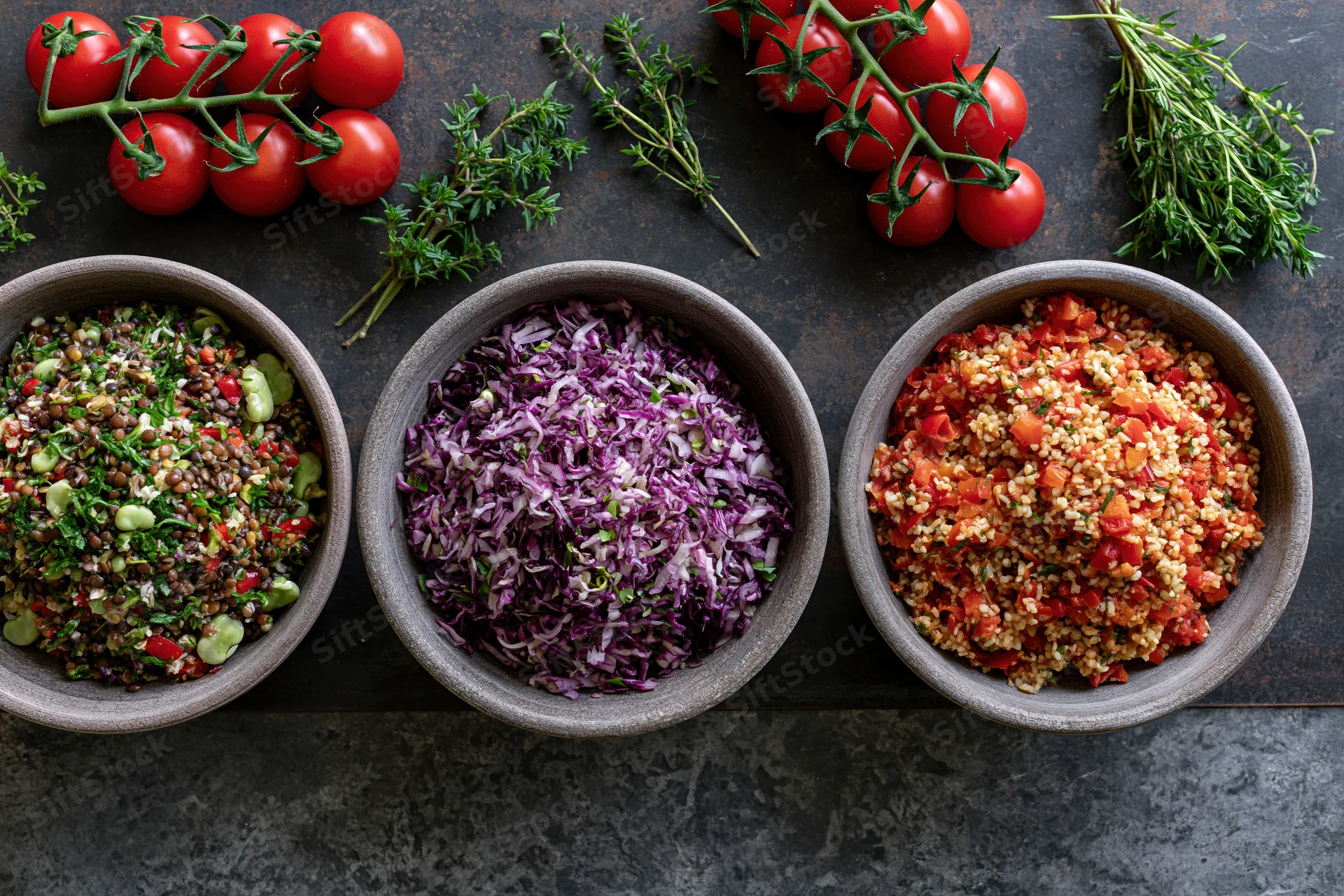 Three rustic bowls of grain and vegetable salads arranged on a dark surface with fresh herbs and tomatoes