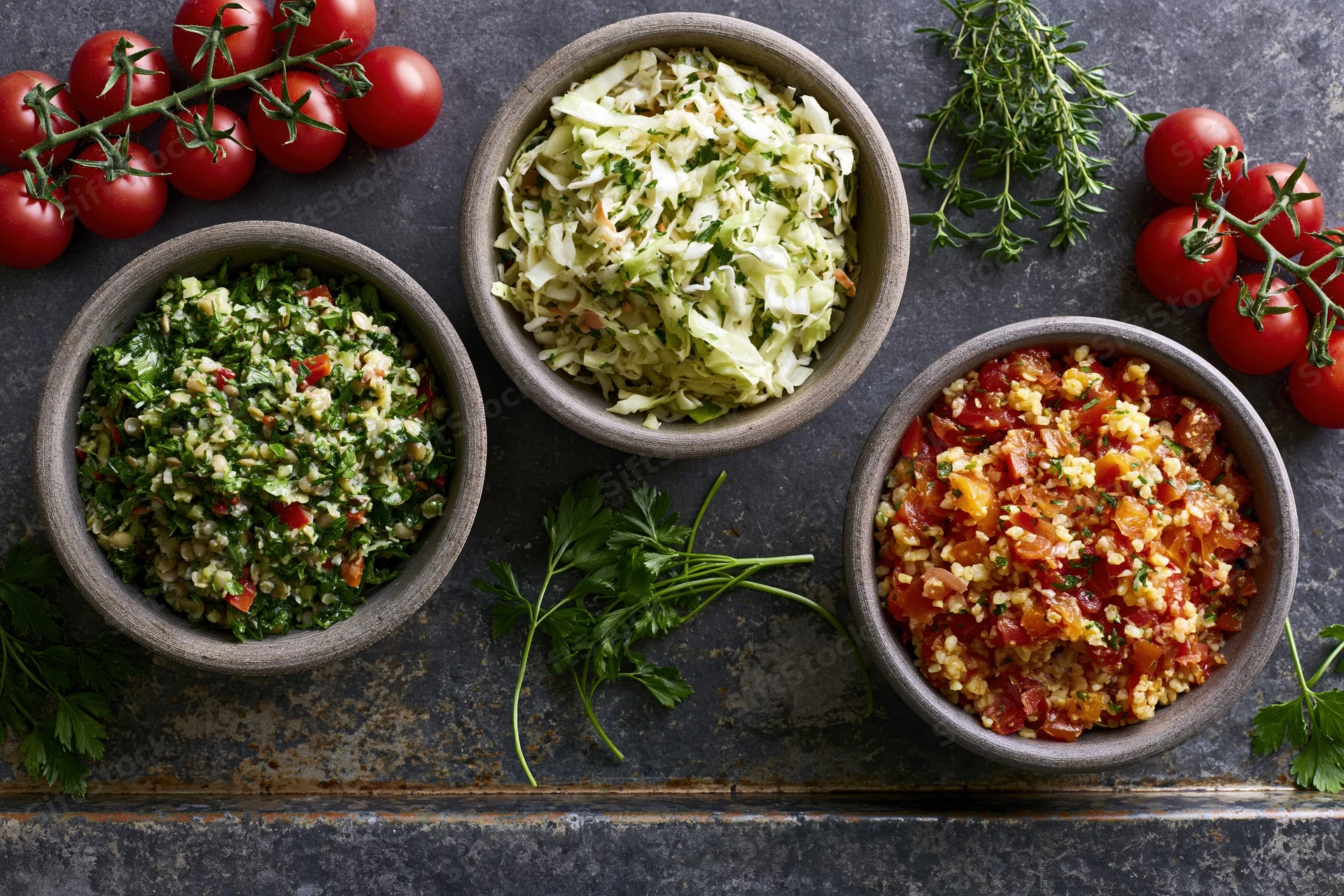 Three rustic bowls of grain and vegetable salads arranged on a dark stone surface with fresh herbs and tomatoes