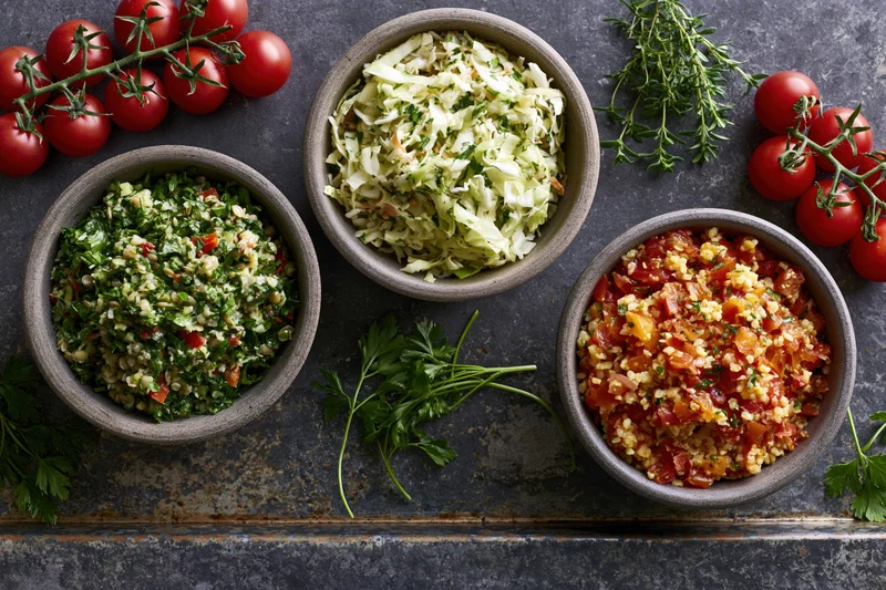 Three rustic bowls of grain and vegetable salads arranged on a dark stone surface with fresh herbs and tomatoes