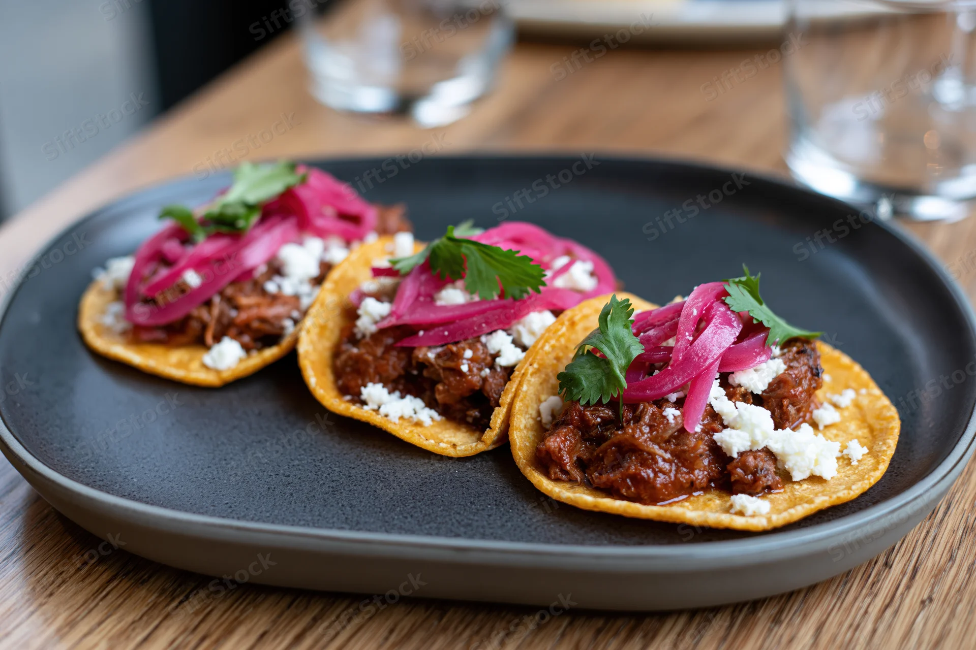 Three small street-style tacos on corn tortillas lined up on a black plate on a wooden table