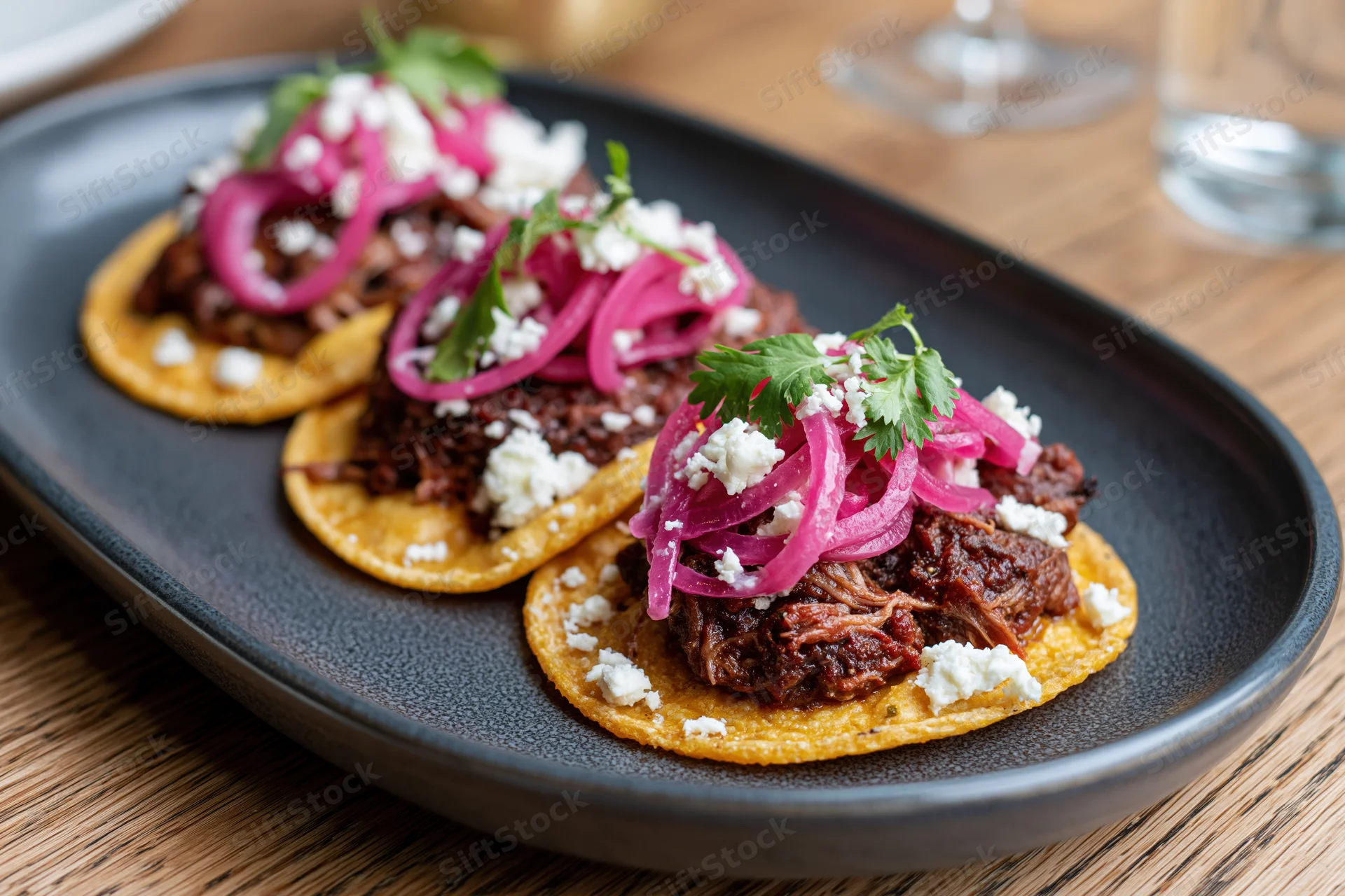 Three street-style tacos with shredded beef, pickled red onions, cilantro and cotija cheese on corn tortillas arranged on a black oval plate on a wooden table