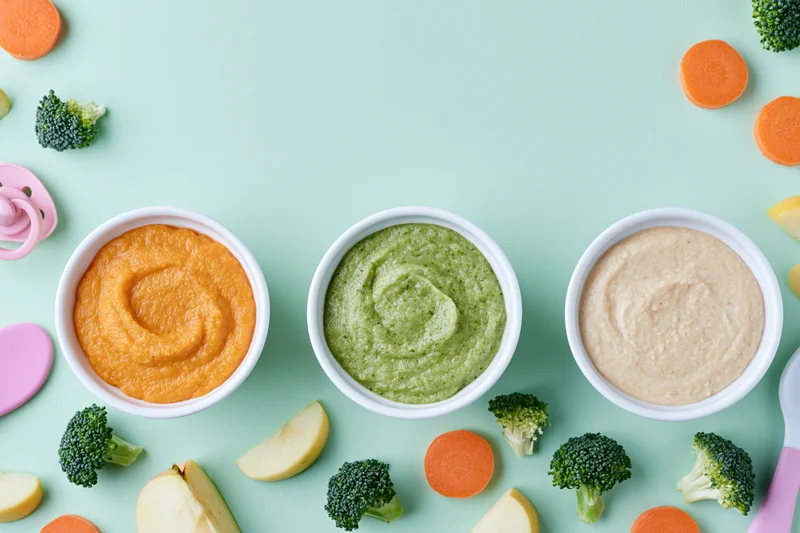 Three bowls of baby food puree with vegetables and fruit arranged on a mint background