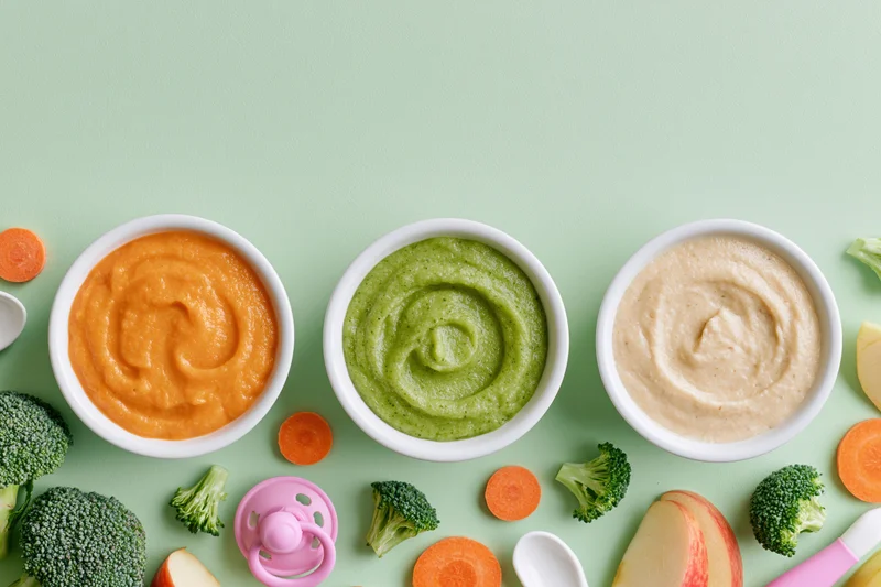 Three bowls of baby food puree with vegetables and fruit arranged on a green surface