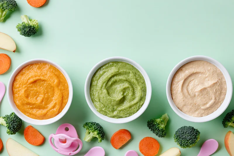 Three white bowls of baby food purees arranged with vegetables and baby items on a mint green surface