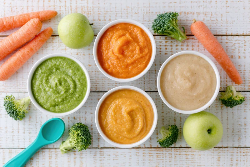 Four bowls of baby food puree with fresh vegetables and a spoon on a wooden table