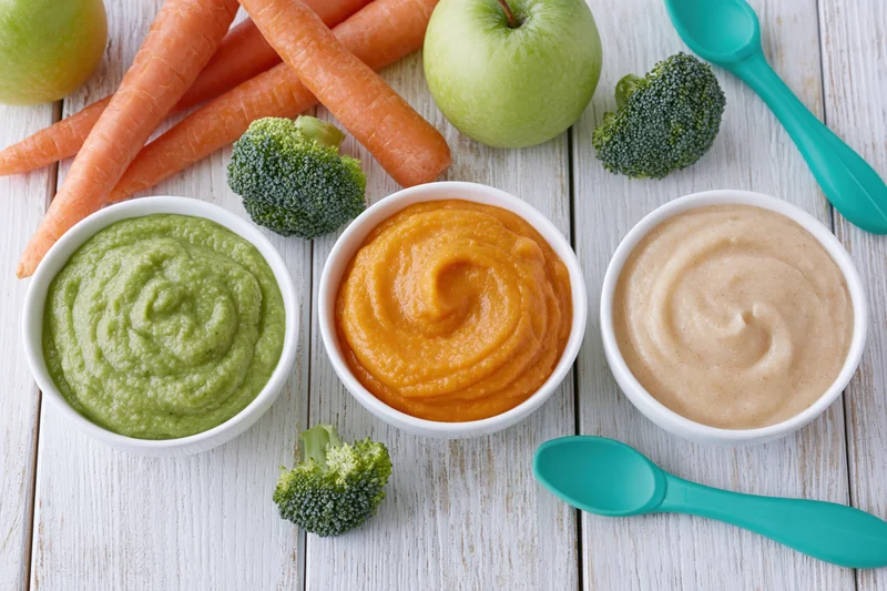 Three white bowls of green, orange and beige baby food puree on a wooden table with fresh vegetables and turquoise spoons