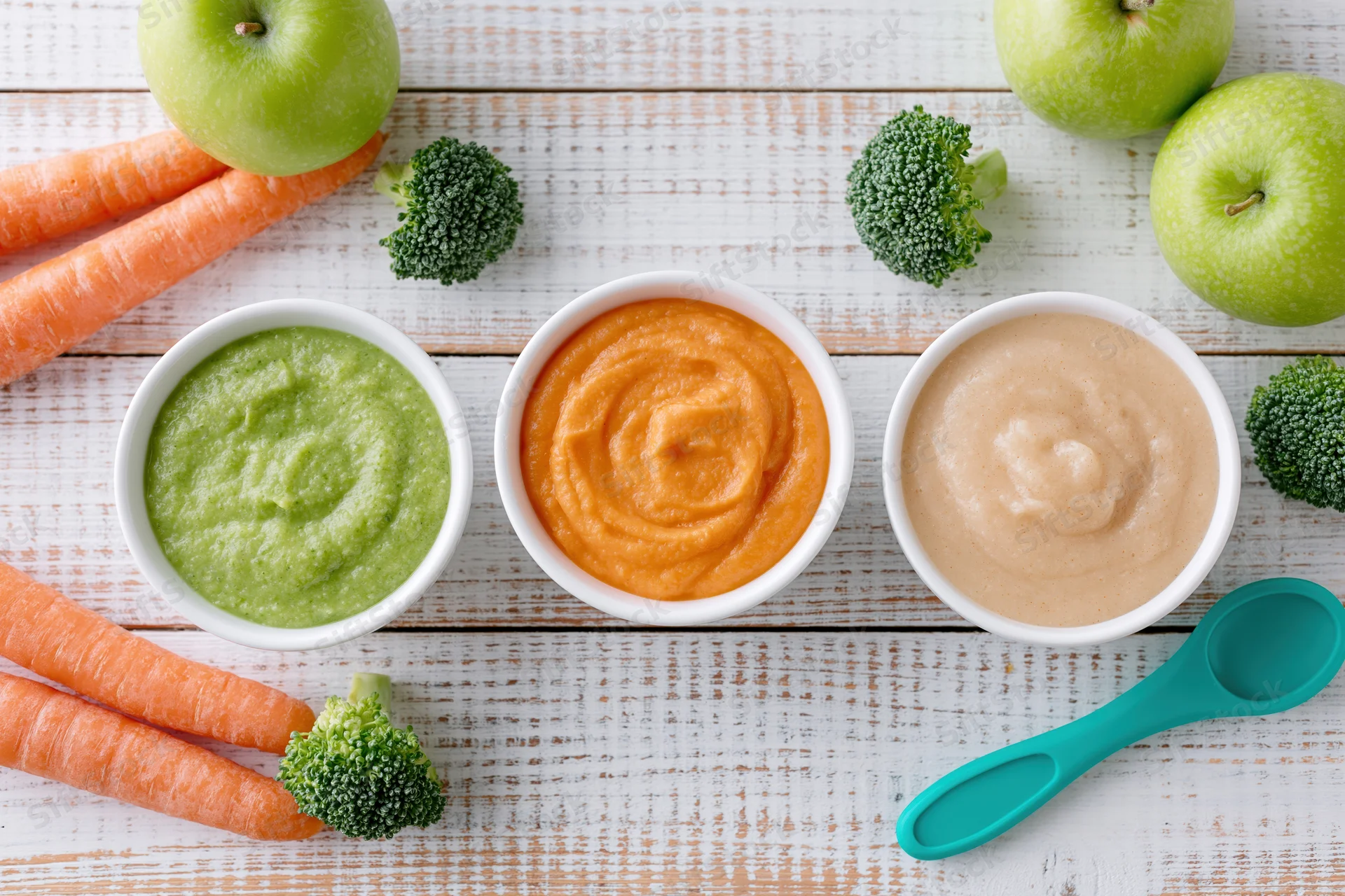 Three white bowls of green, orange and beige baby food puree arranged on a wooden table with fresh carrots, broccoli and apples around them