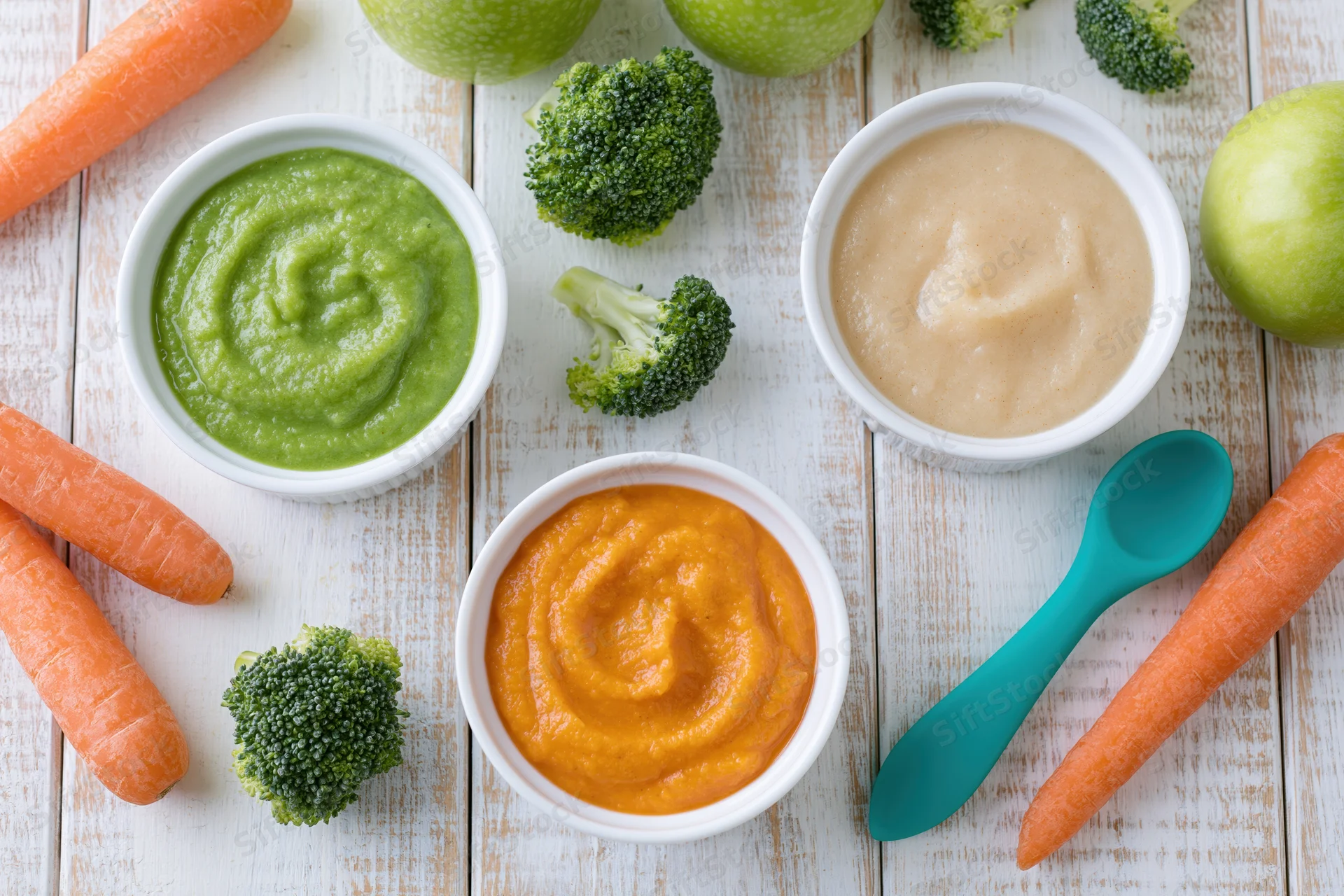 Three white bowls of colorful baby food purees arranged with fresh vegetables on a wooden table