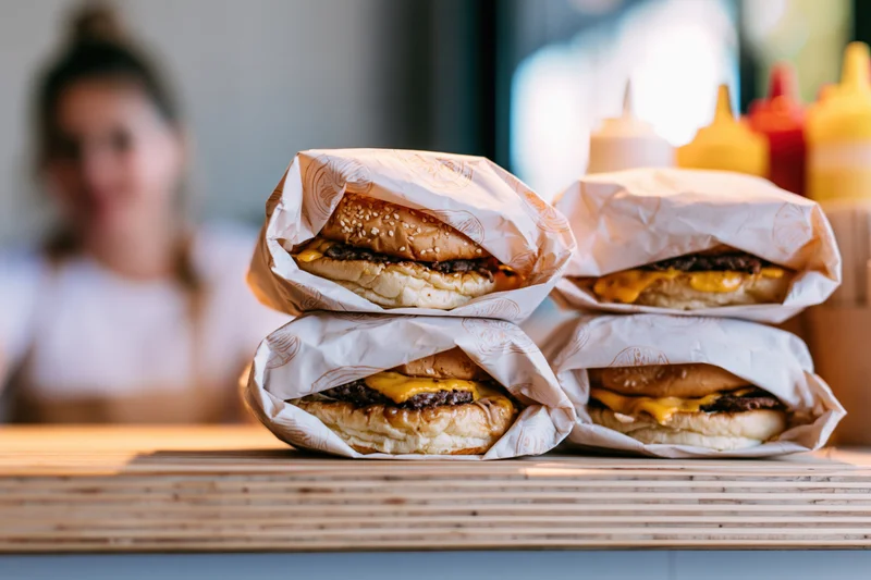 Four cheeseburgers wrapped in parchment paper stacked on a wooden counter with condiment bottles and a blurred woman in the background