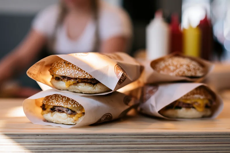 Three cheeseburgers wrapped in branded paper stacked on a wooden table with condiments in background