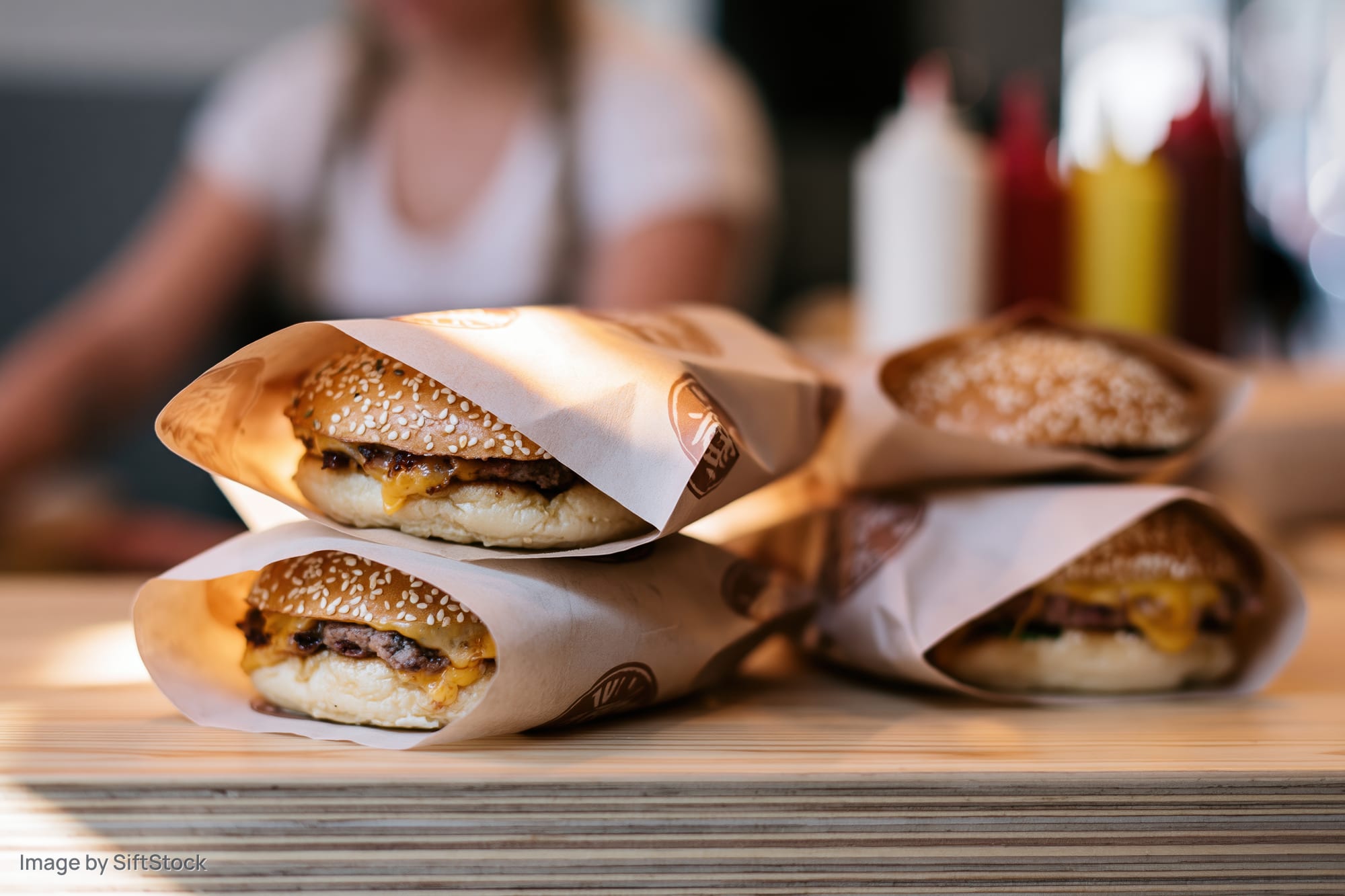 Three cheeseburgers wrapped in branded paper stacked on a wooden table with condiments in background