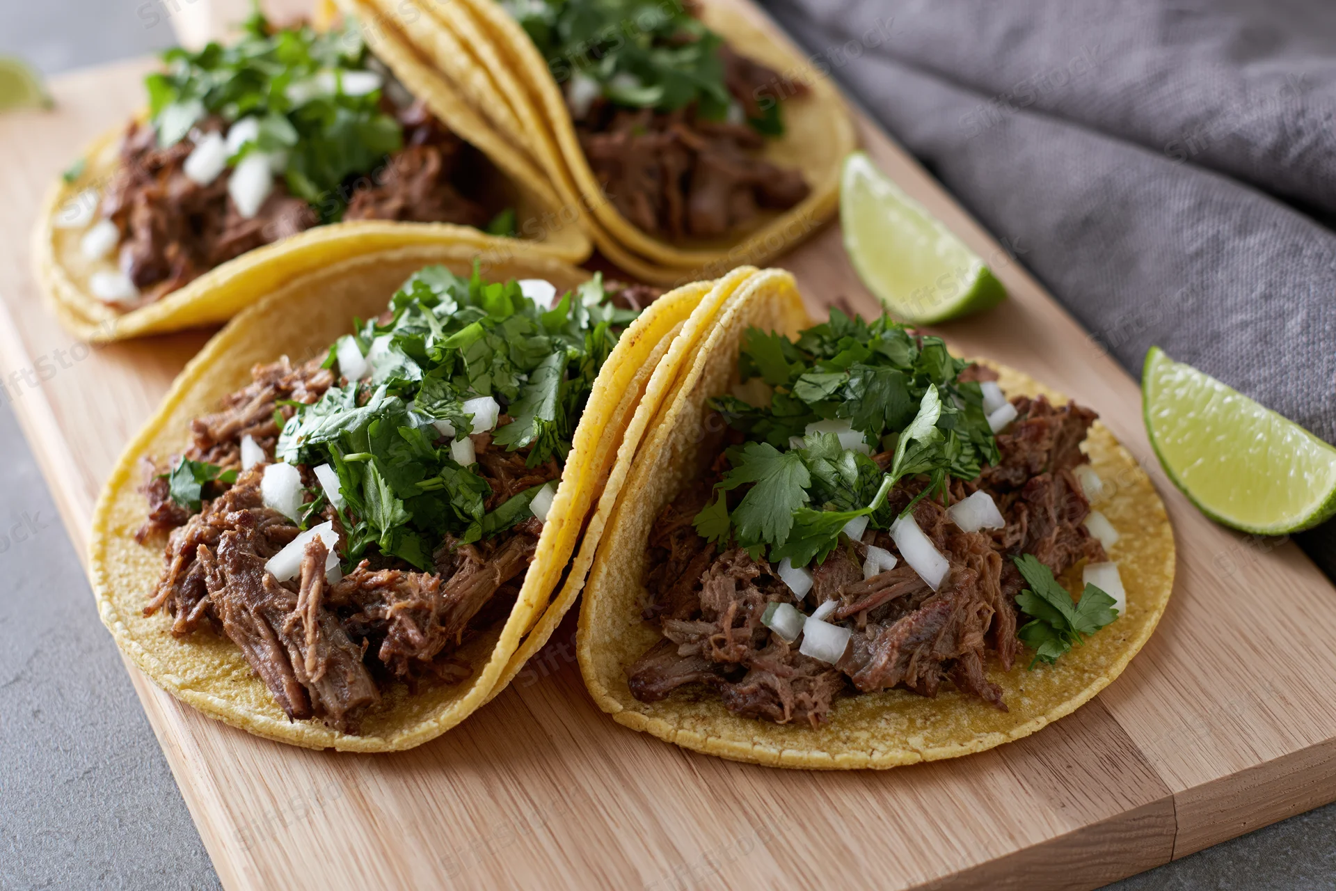 Four street-style tacos with shredded beef, cilantro, onion and lime on a wooden cutting board