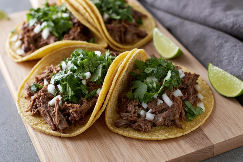 Four street-style tacos with shredded beef, cilantro, onion and lime on a wooden cutting board