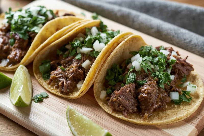 Three street-style tacos with braised beef and fresh toppings on a wooden cutting board