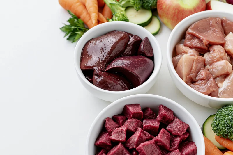 Raw chicken, liver, and beef in white ceramic bowls with fresh vegetables arranged on a white surface